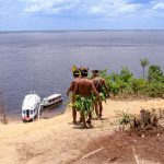 Indigenous men in traditional attire overlooking boats on the Amazon River.